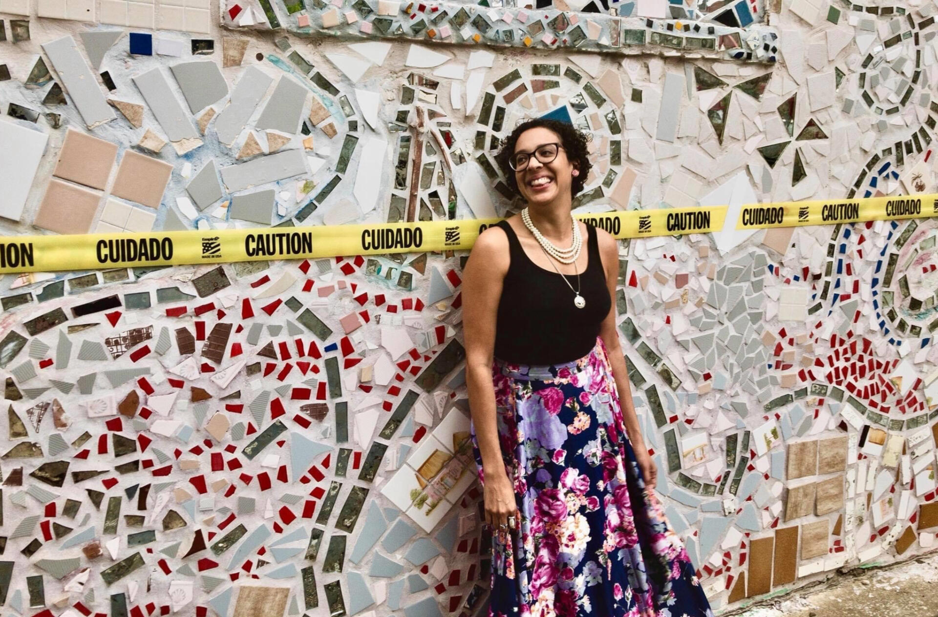 Photograph of Lexi Jones, the artist, wearing a floral skirt, black tank top, pearls and glasses.